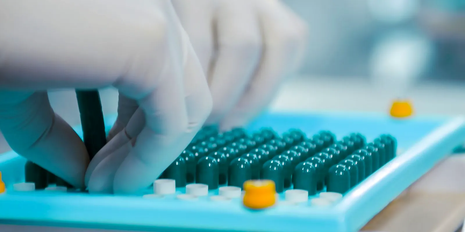 Mano con guante cargando cápsulas en una placa de llenado en un laboratorio farmacéutico